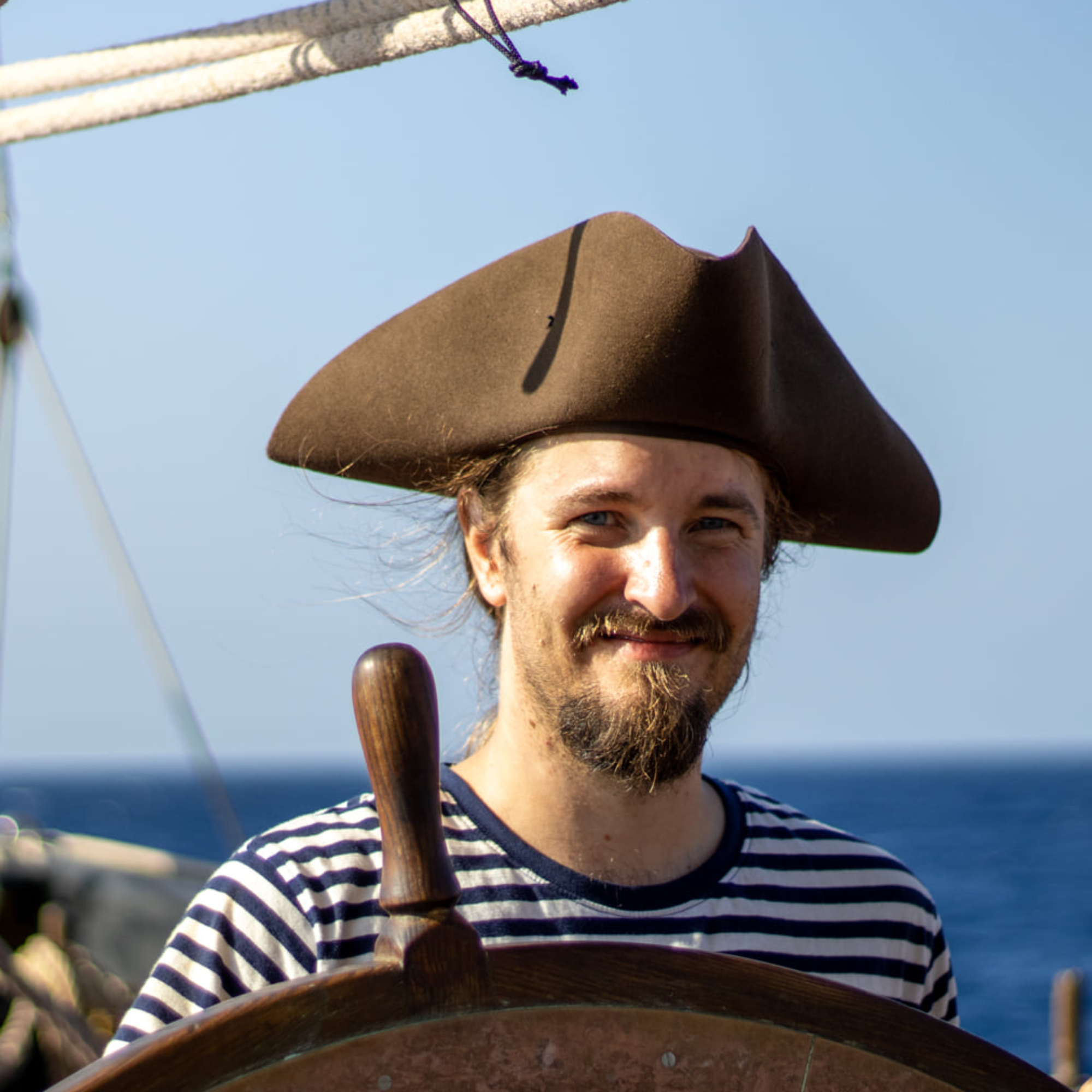 my portrait - a man with blue eyes and long hair, in his thirties, in a pirate hat and striped shirt, behind a wheel.